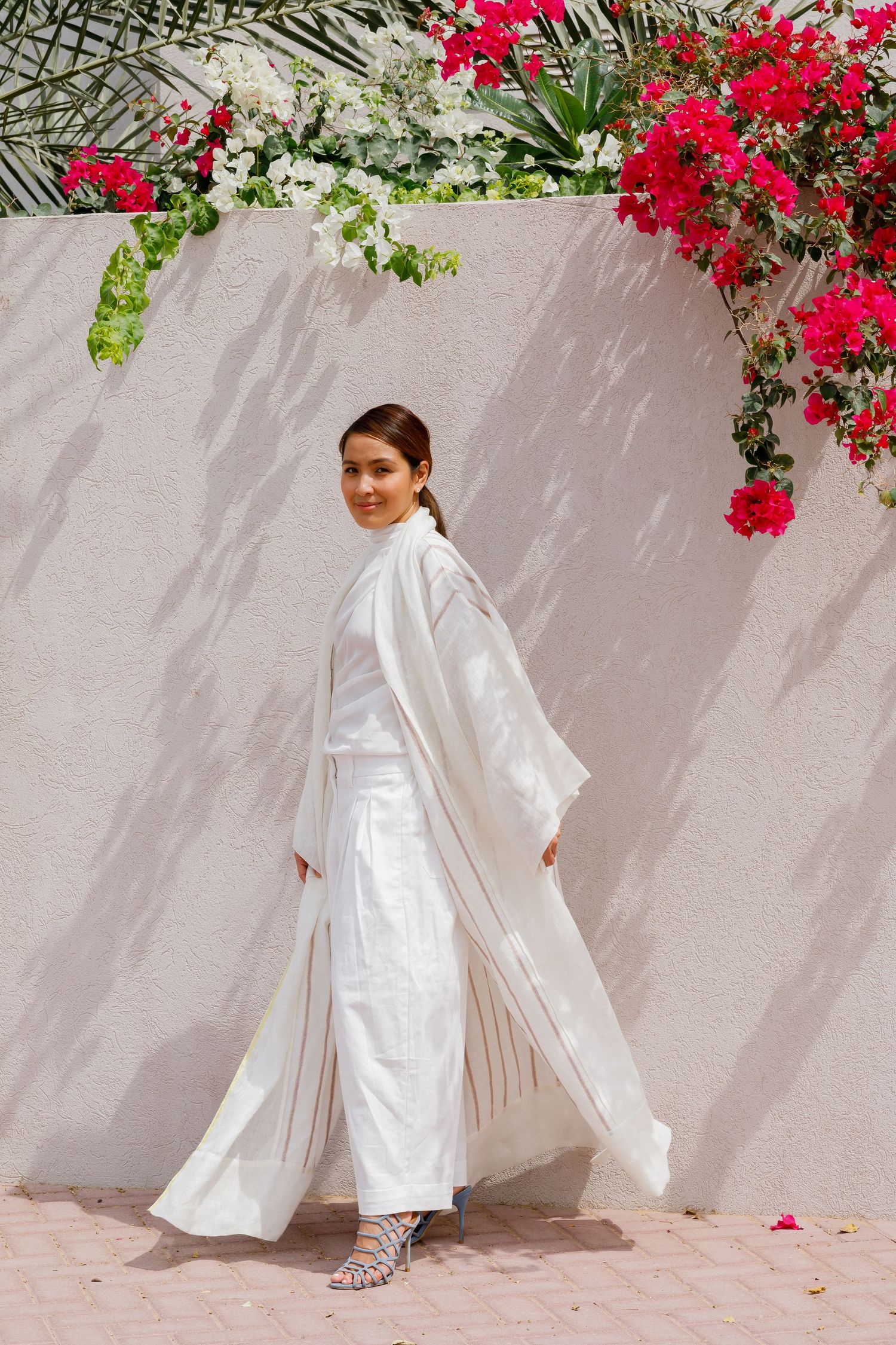 A figure in an all-white flowing ensemble stands against a blush wall adorned with vibrant red bougainvillea flowers.