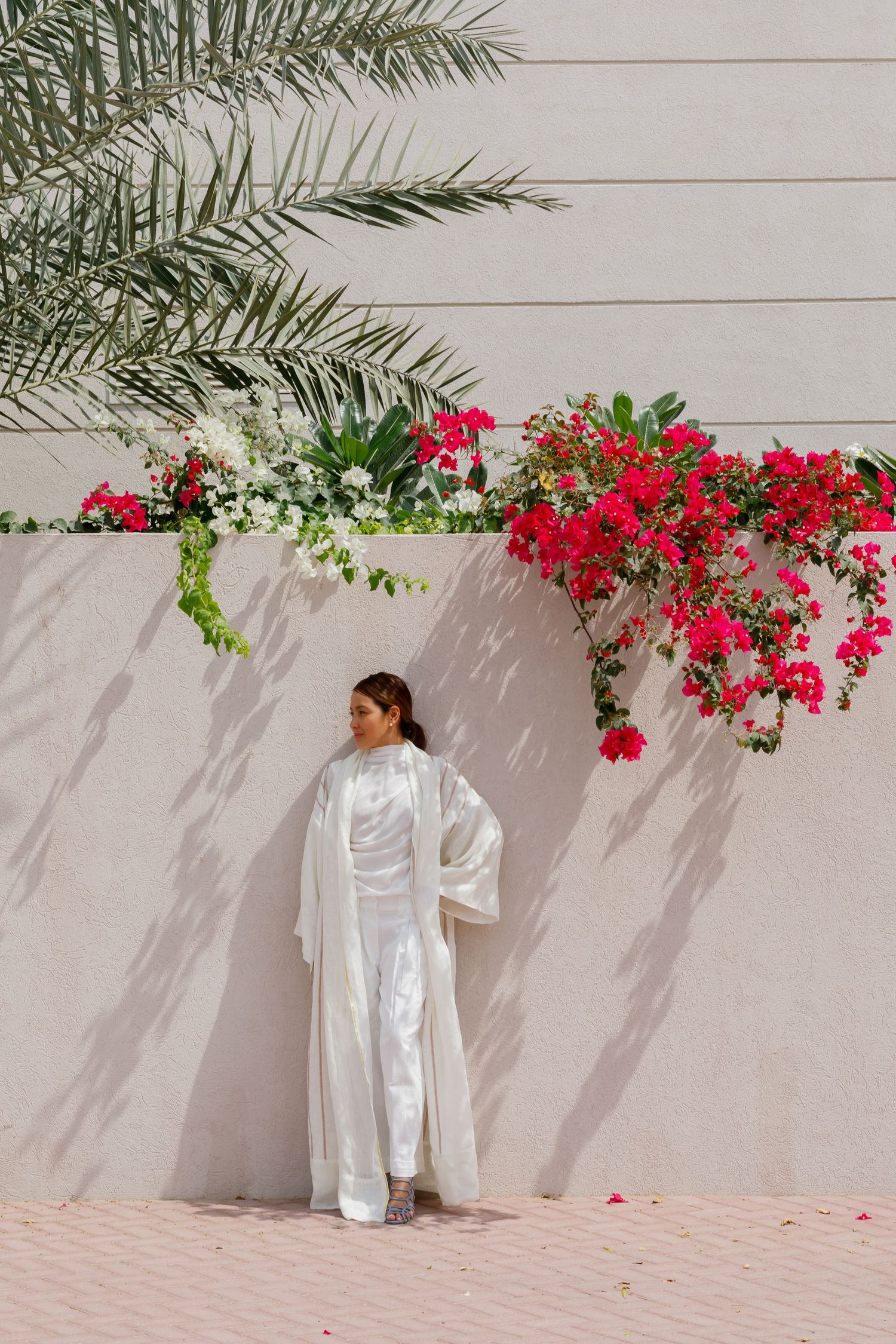 Figure in flowing white outfit stands against light wall with vibrant pink bougainvillea flowers and palm fronds above.