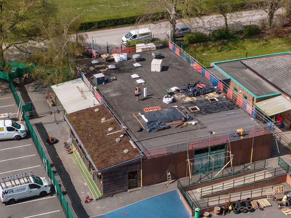 Aerial view of a commercial strip mall parking lot with vehicles and buildings surrounded by green landscaping.