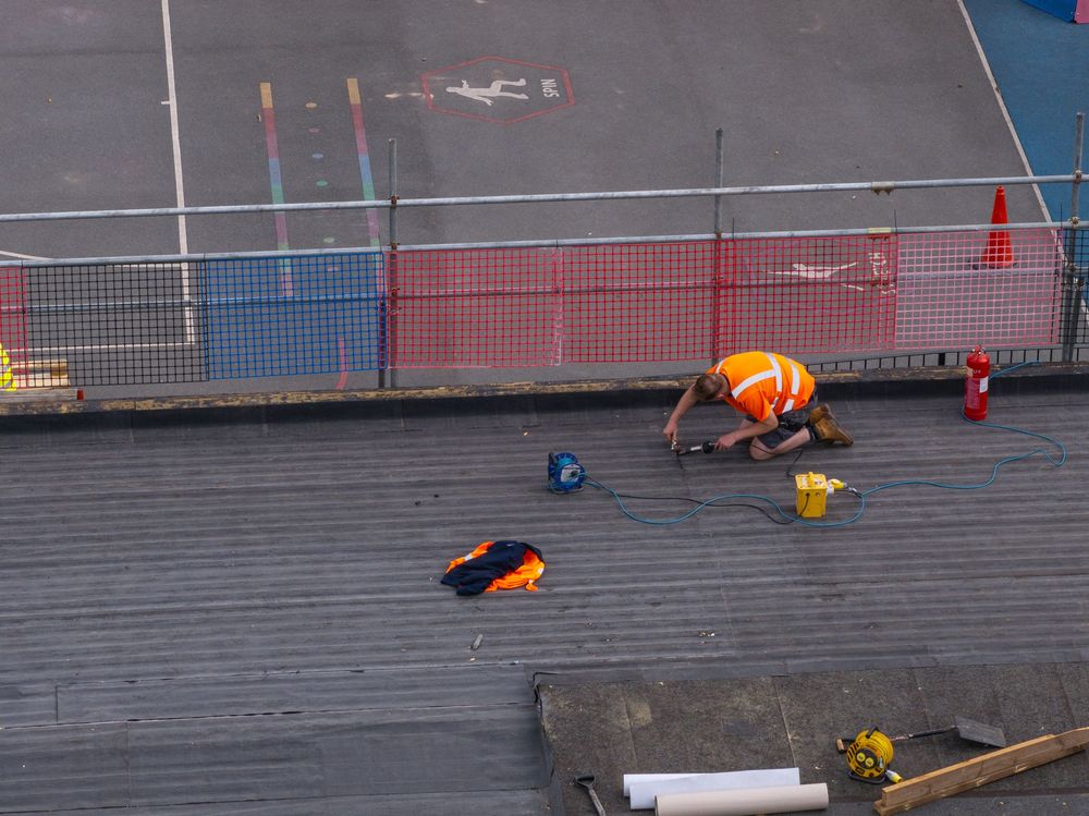 Construction workers in orange safety gear working on a flat roof surface next to protective barriers.