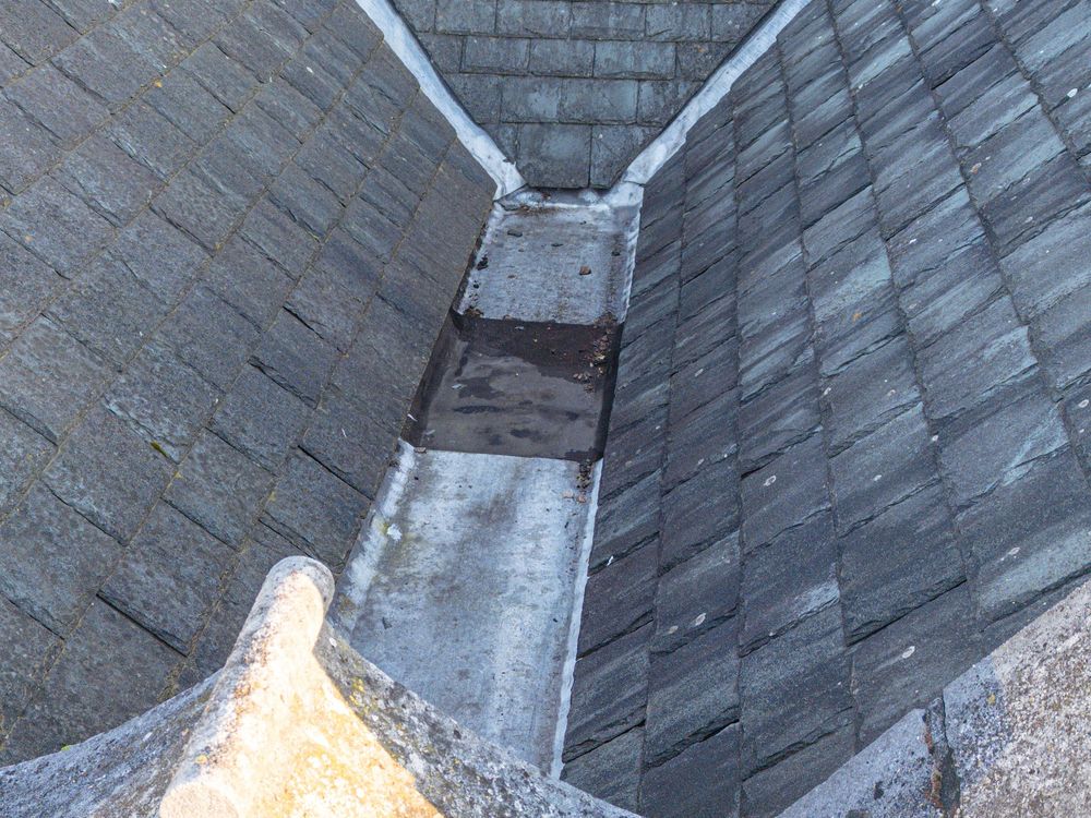 A close-up view of a residential roof valley showing slate shingles and a metal lined drainage channel.