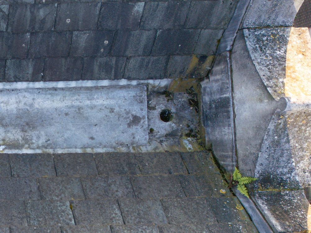 Close-up view of a roof corner showing weathered slate tiles and moss growth along metal flashing detail.