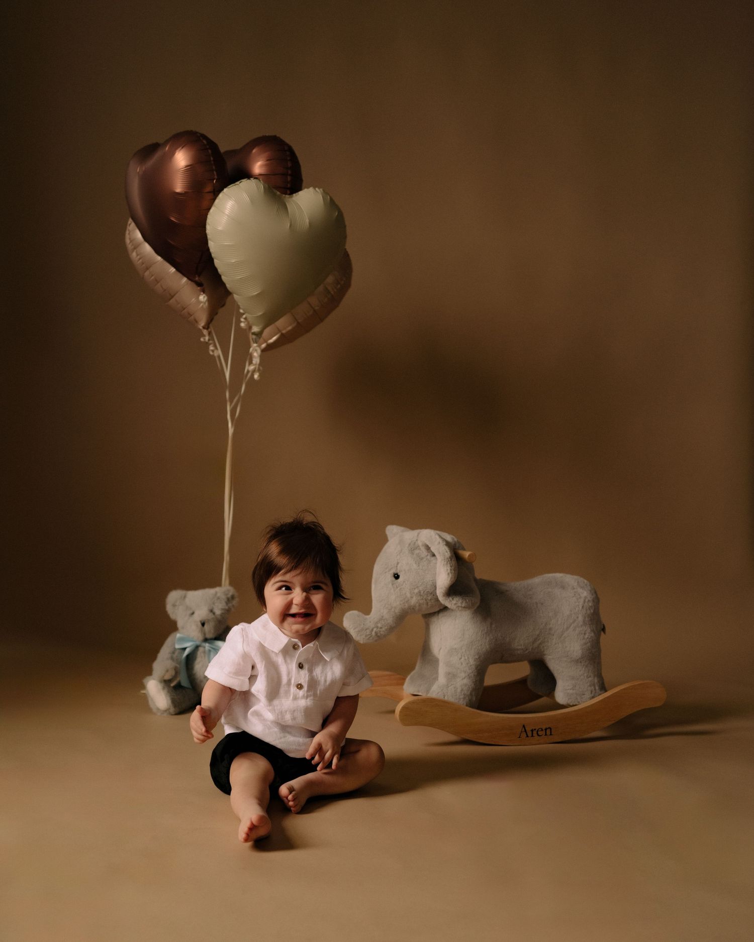 Baby sits on brown backdrop with heart balloons and rocking horse toy in studio setting.
