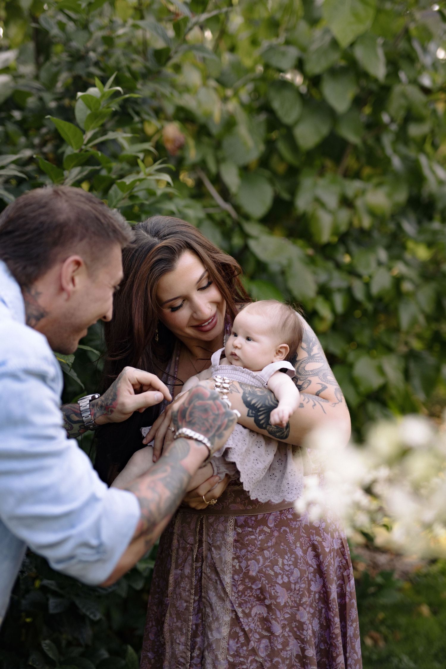 A couple shares a tender moment with their baby outdoors in a garden setting with green foliage in the background.