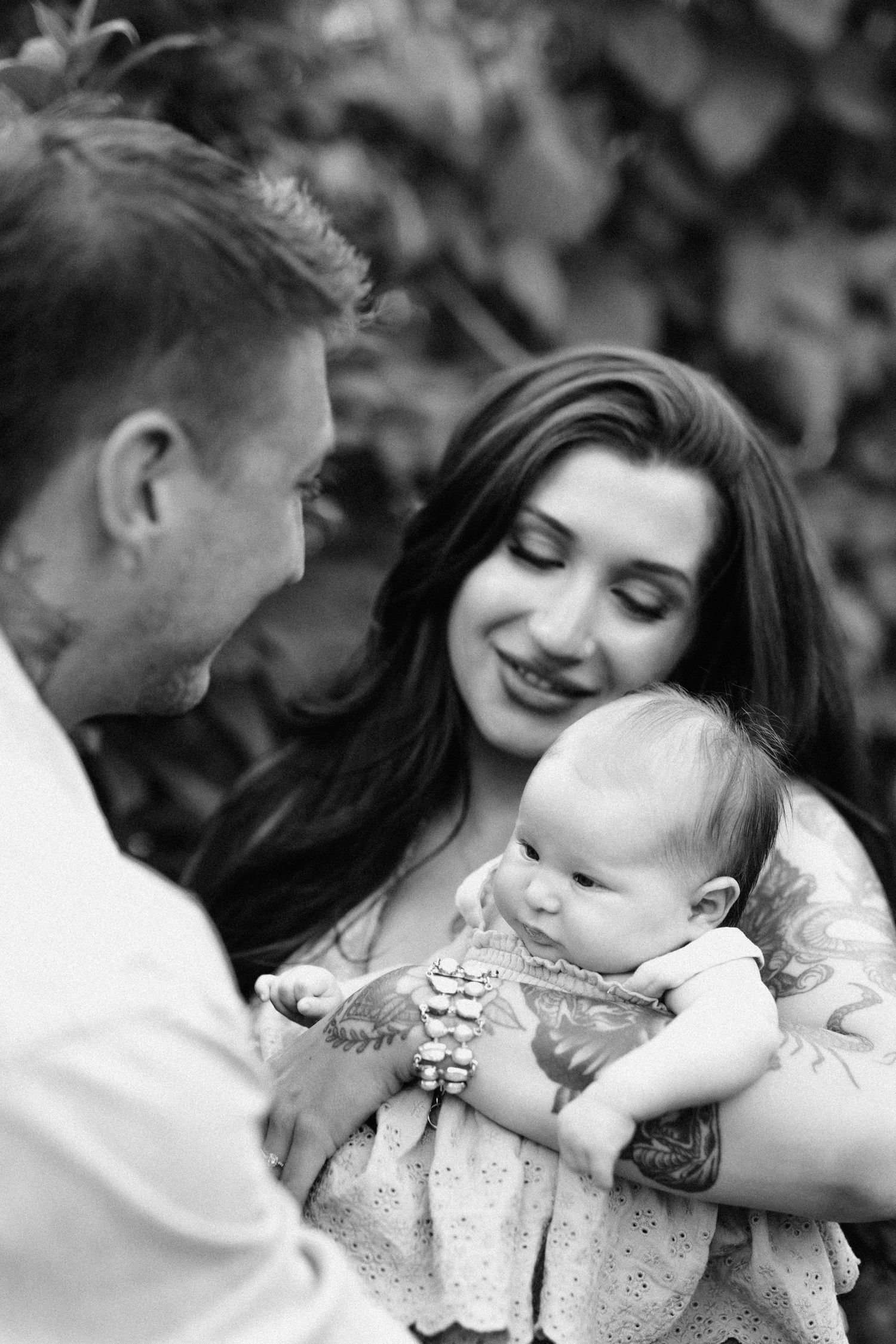 A tender black and white family moment with parents gazing lovingly at their baby in a floral dress.