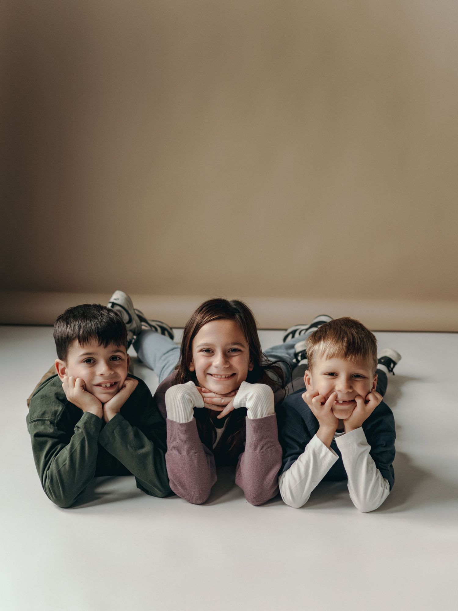 Three children lying side by side on a light colored floor, resting their heads on their hands and smiling at the camera.