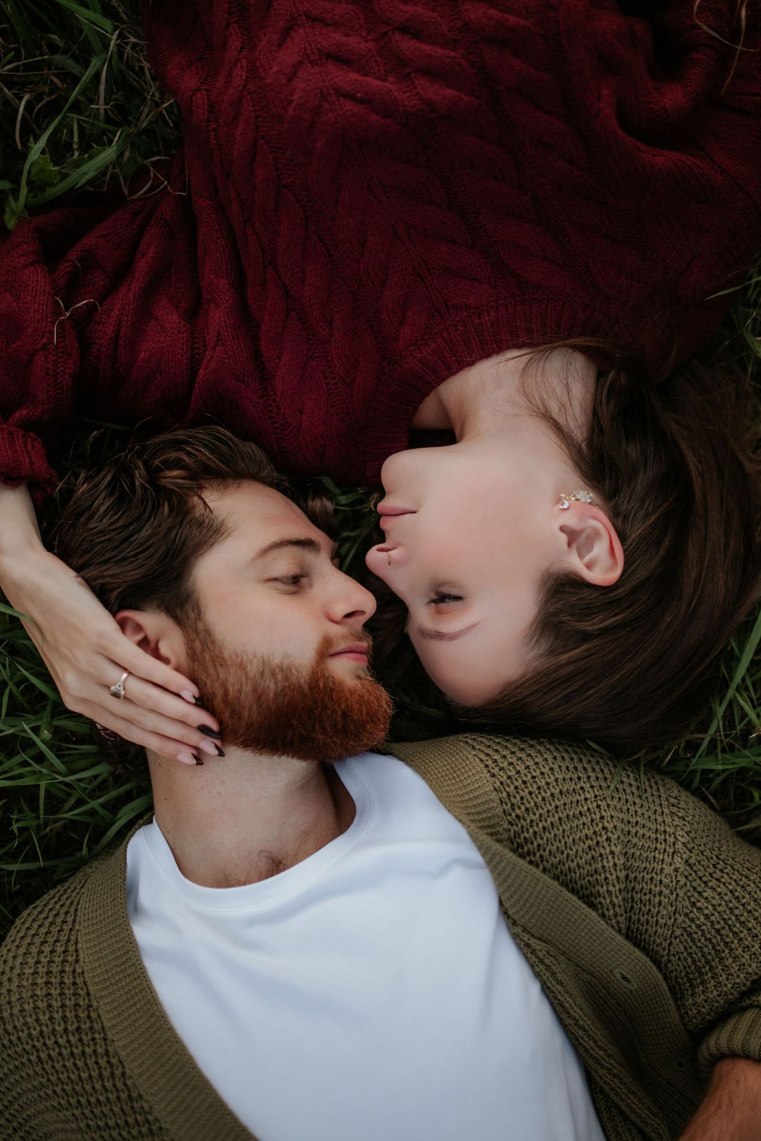 Couple lying head to head on green grass, a bearded man in an olive cardigan and a woman in a burgundy sweater leaning in for a kiss.