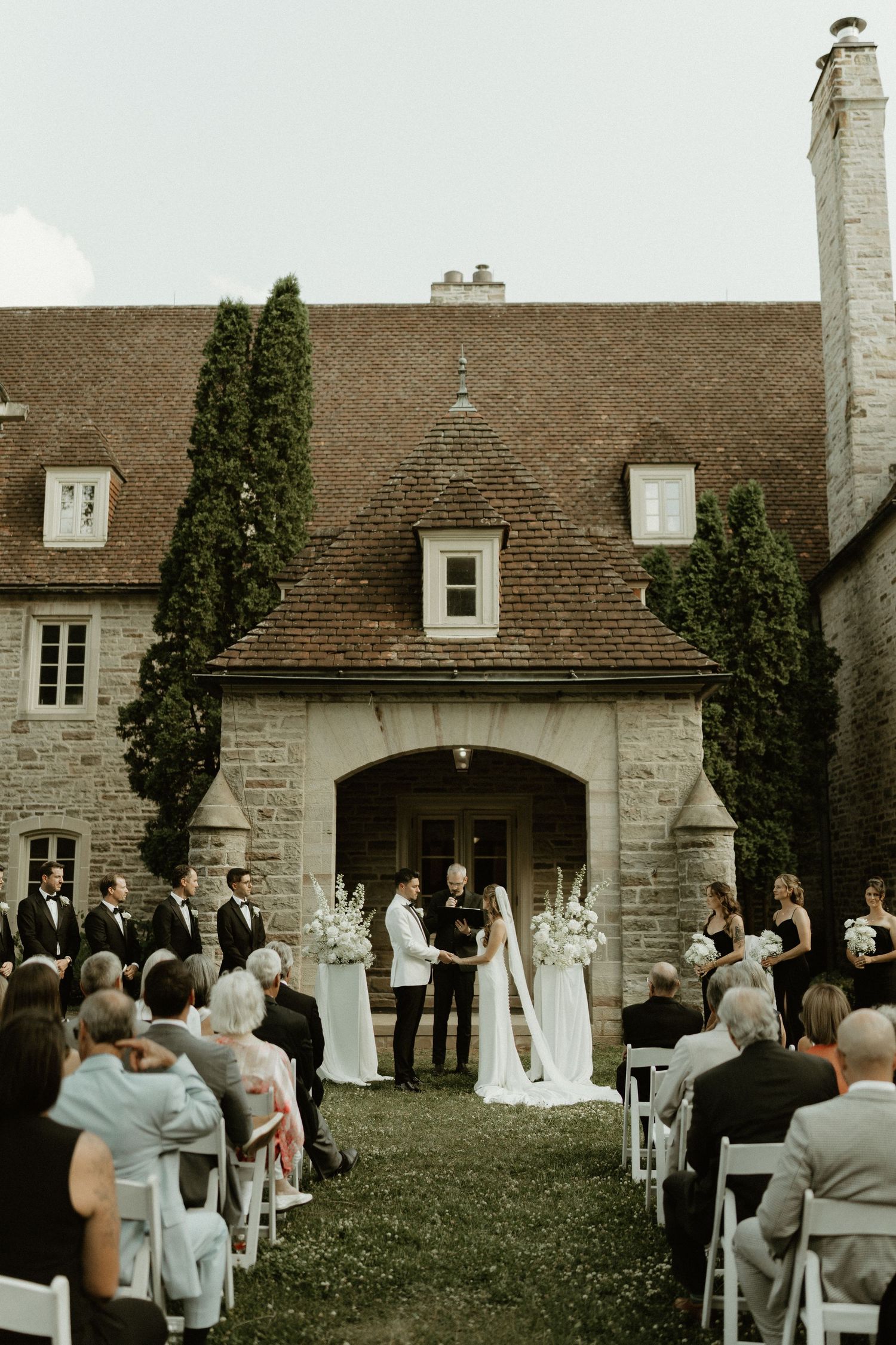 An outdoor wedding ceremony takes place in front of a historic stone mansion with arched doorway and climbing ivy.