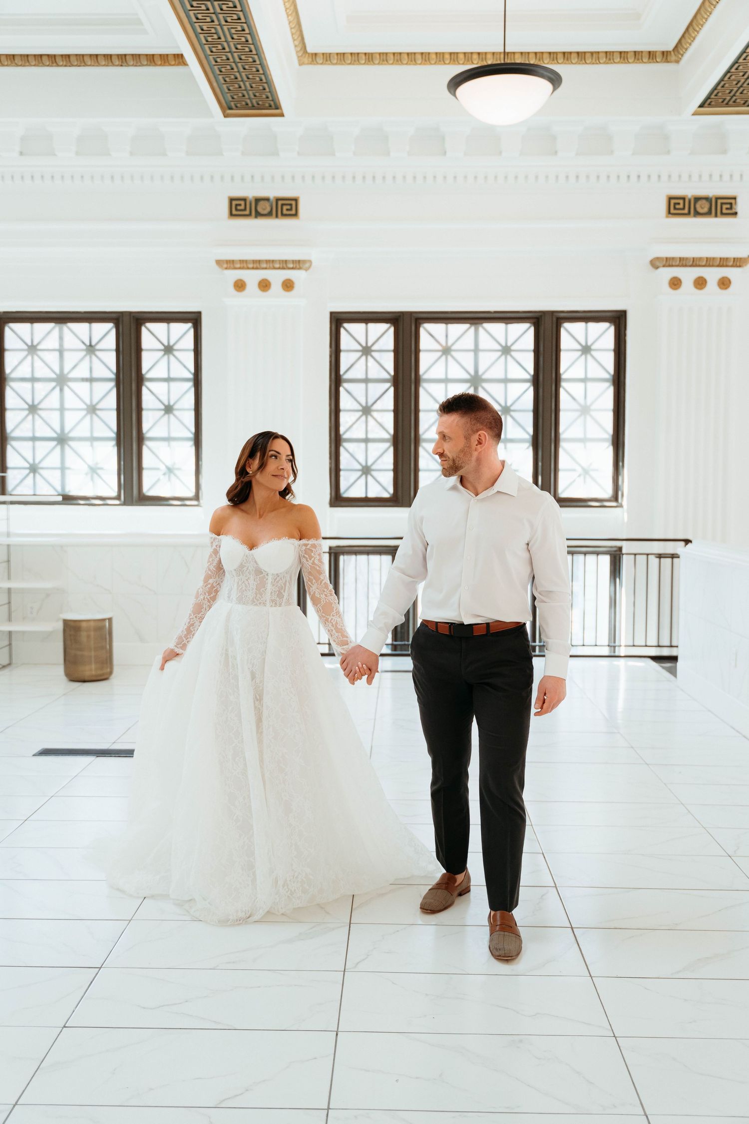 Couple in wedding attire dancing in bright white room with ornate windows.