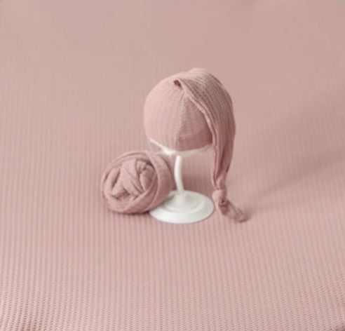 Pink knitted newborn photography props including hat and wrap on rose colored background.