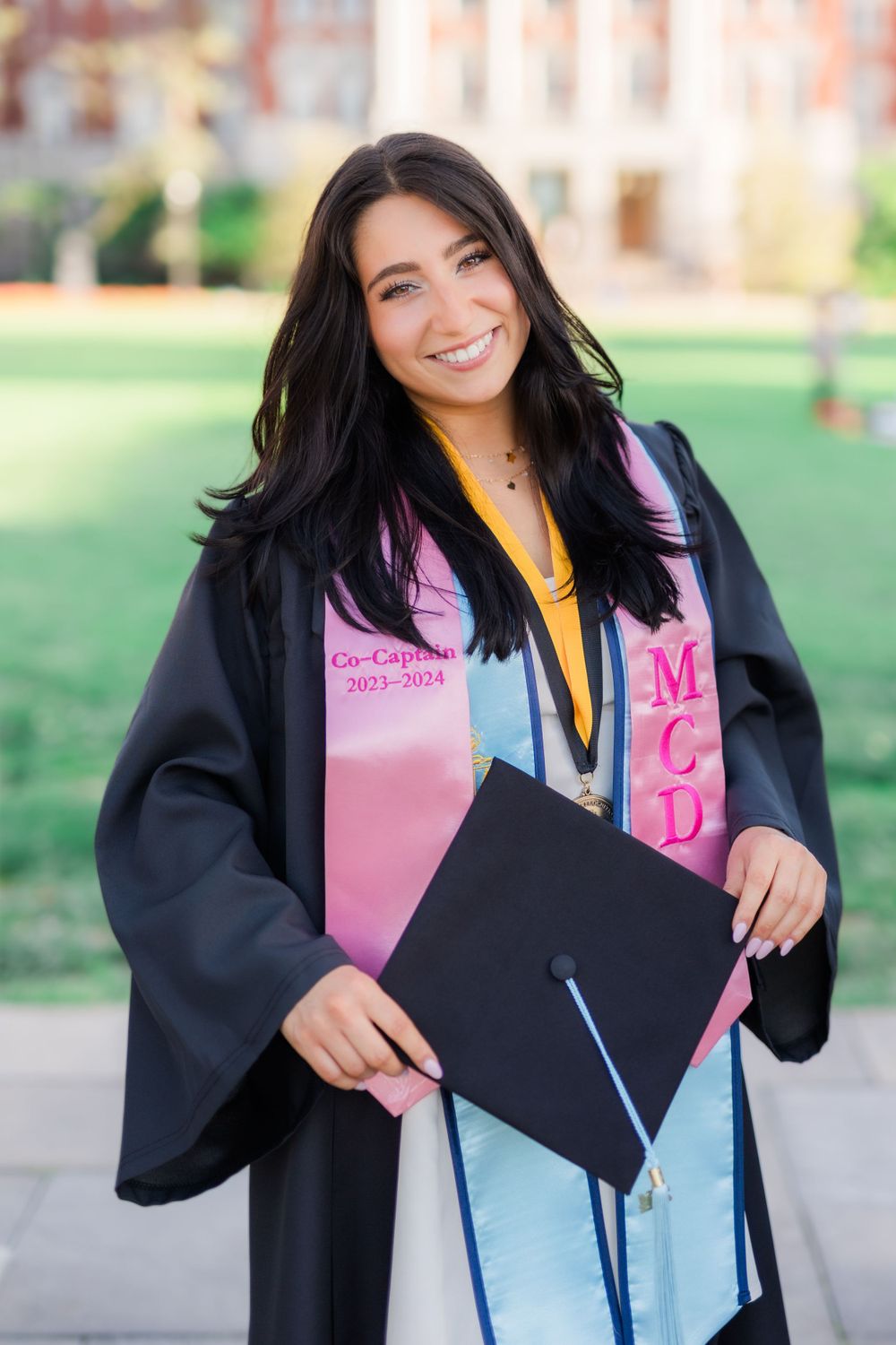 Graduate in black gown with pink sash holds diploma while standing on campus lawn.