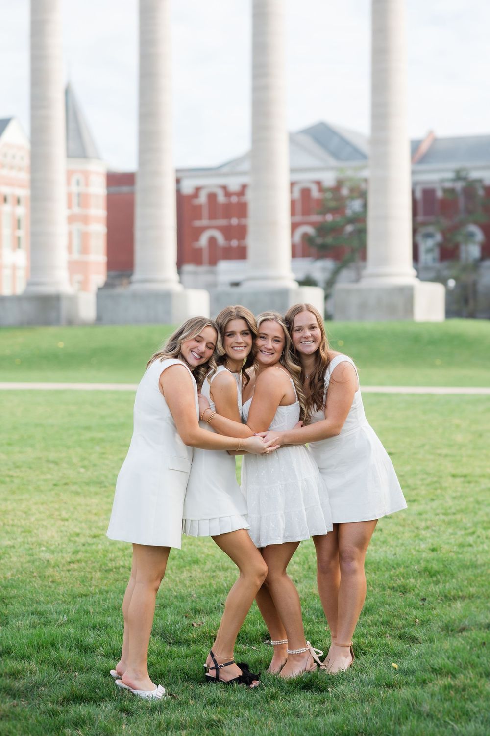A group of friends wearing white dresses huddle together and smile on a grassy lawn in front of white columns.