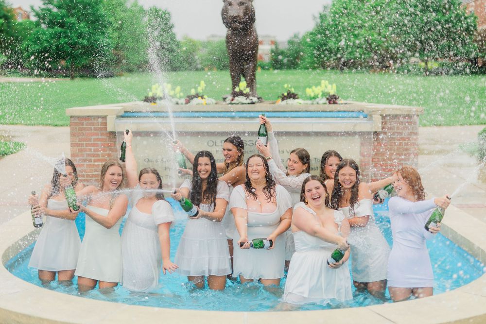 Group of bridesmaids in light blue dresses posing together by a fountain statue.