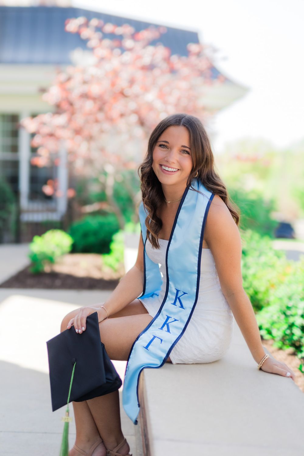 Graduate in white dress and light blue sash sits outdoors with graduation cap near flowering tree.