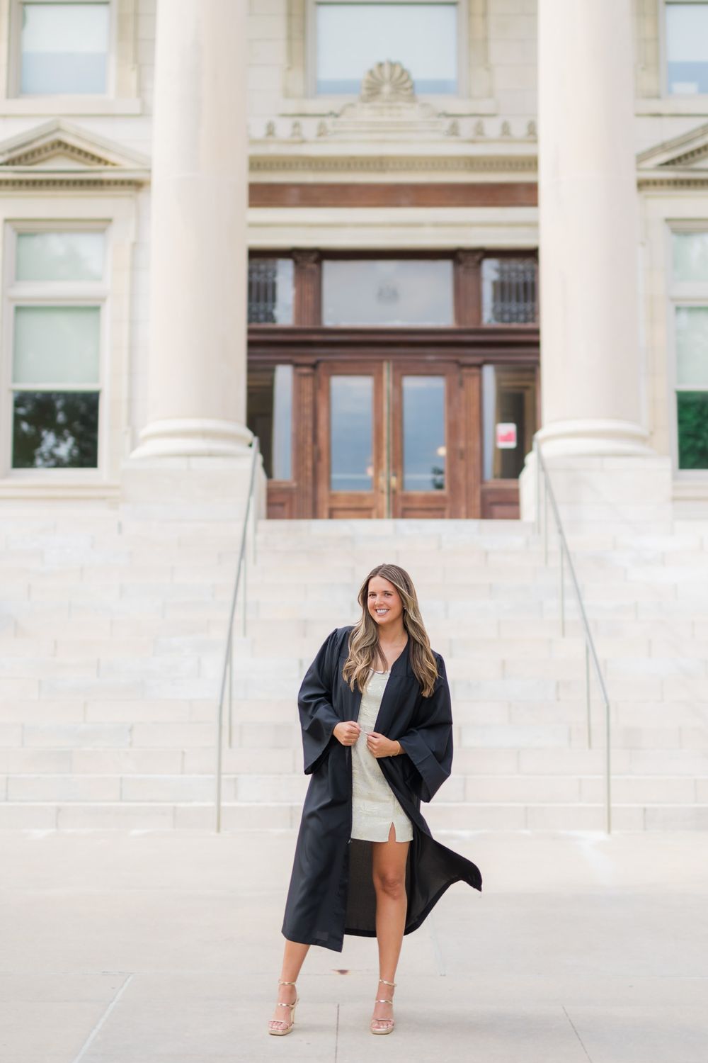 Graduate in black cap and gown poses on white stone steps of columned building entrance.