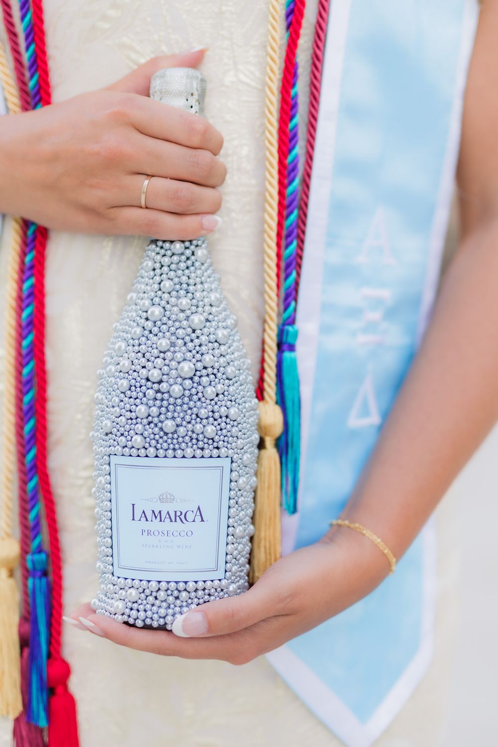 Decorative bottle of Lamarca prosecco covered in silver beads being held against a colorful braided rope background.