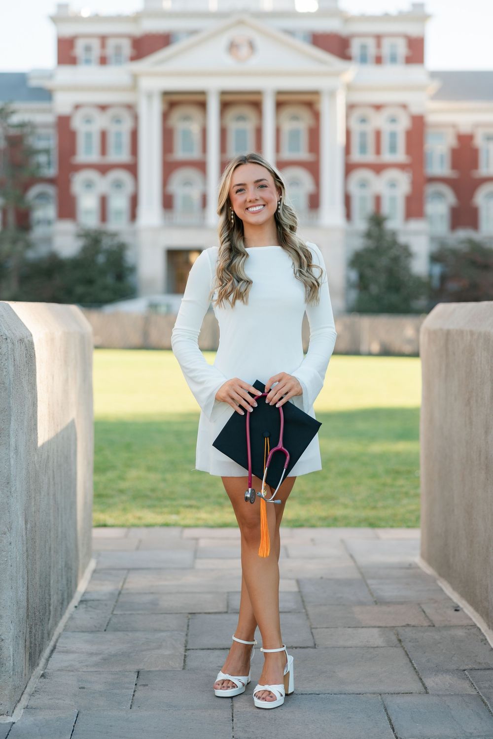 A graduate in a white bell sleeve dress poses with their diploma in front of a grand brick university building.