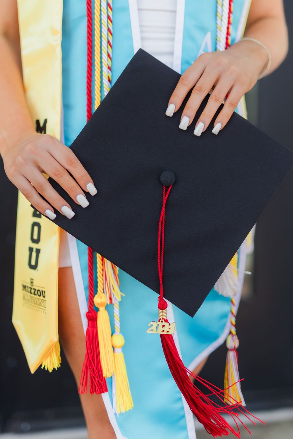 Hands holding a black graduation cap with red tassel against colorful graduation stoles and cords.