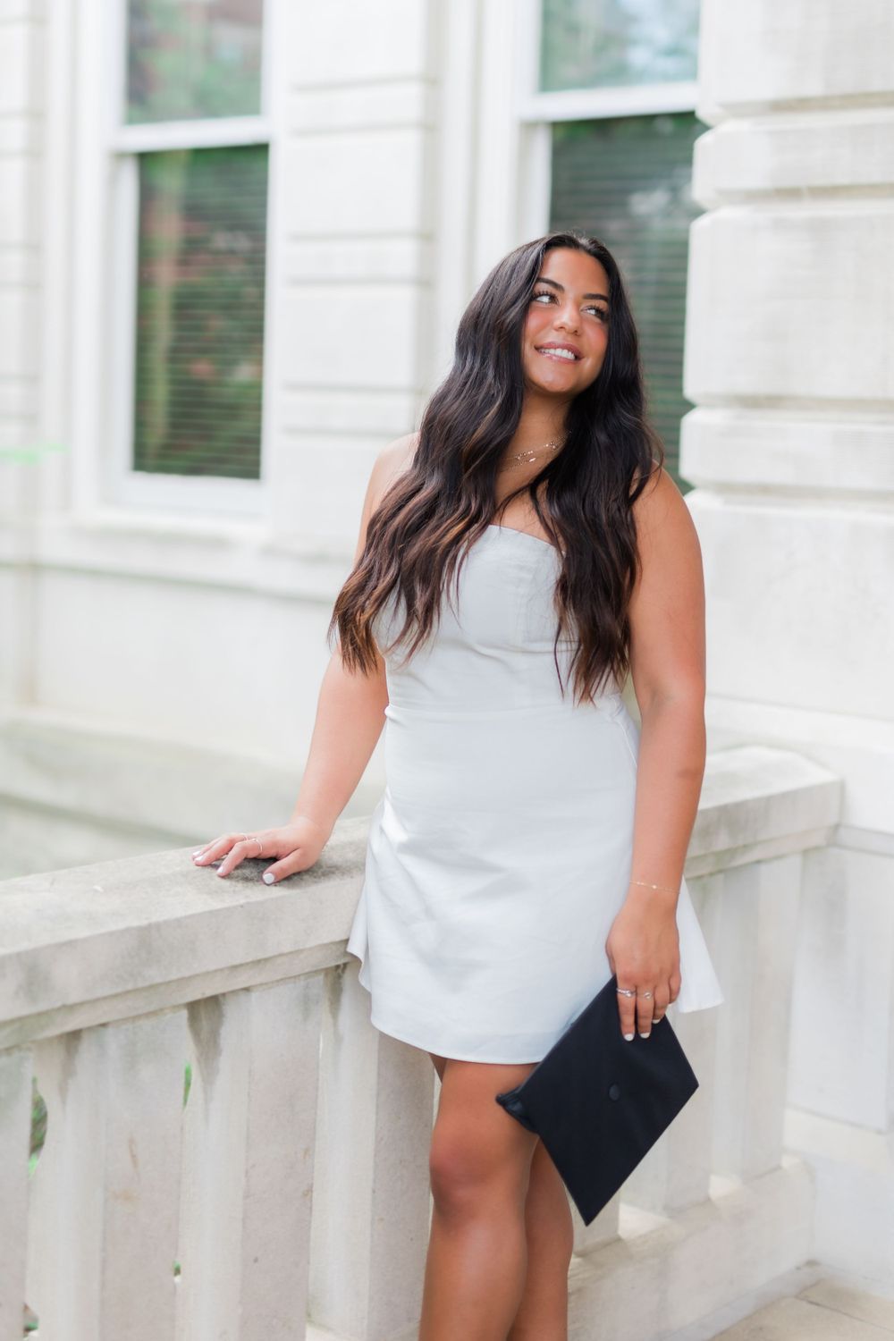 Graduate in white dress holds black graduation cap while posing against white building exterior.