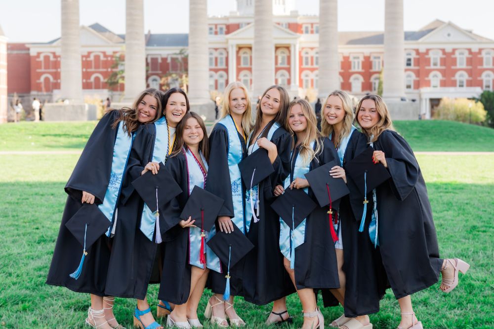 Graduates in black caps and gowns with blue sashes pose together on campus lawn.