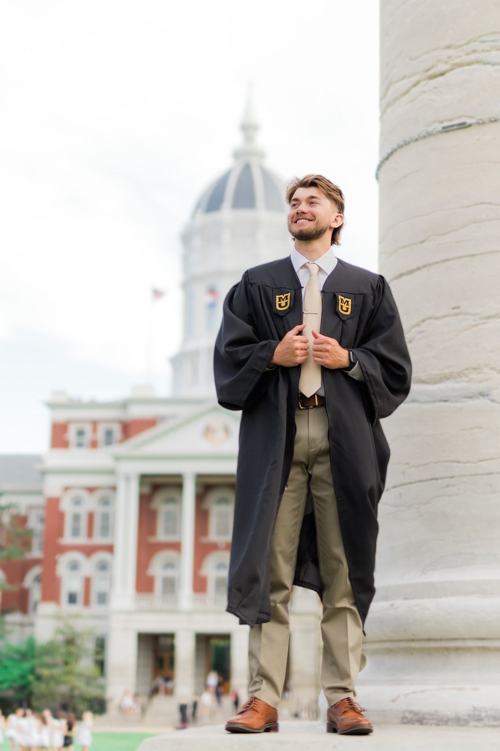 Graduate in black cap and gown stands by white column with domed building in background.