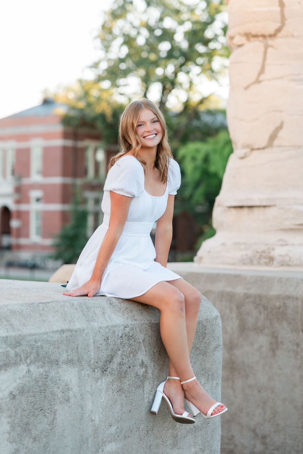 Someone in a short white dress sits on a concrete ledge wearing strappy heels with a brick building in the background.