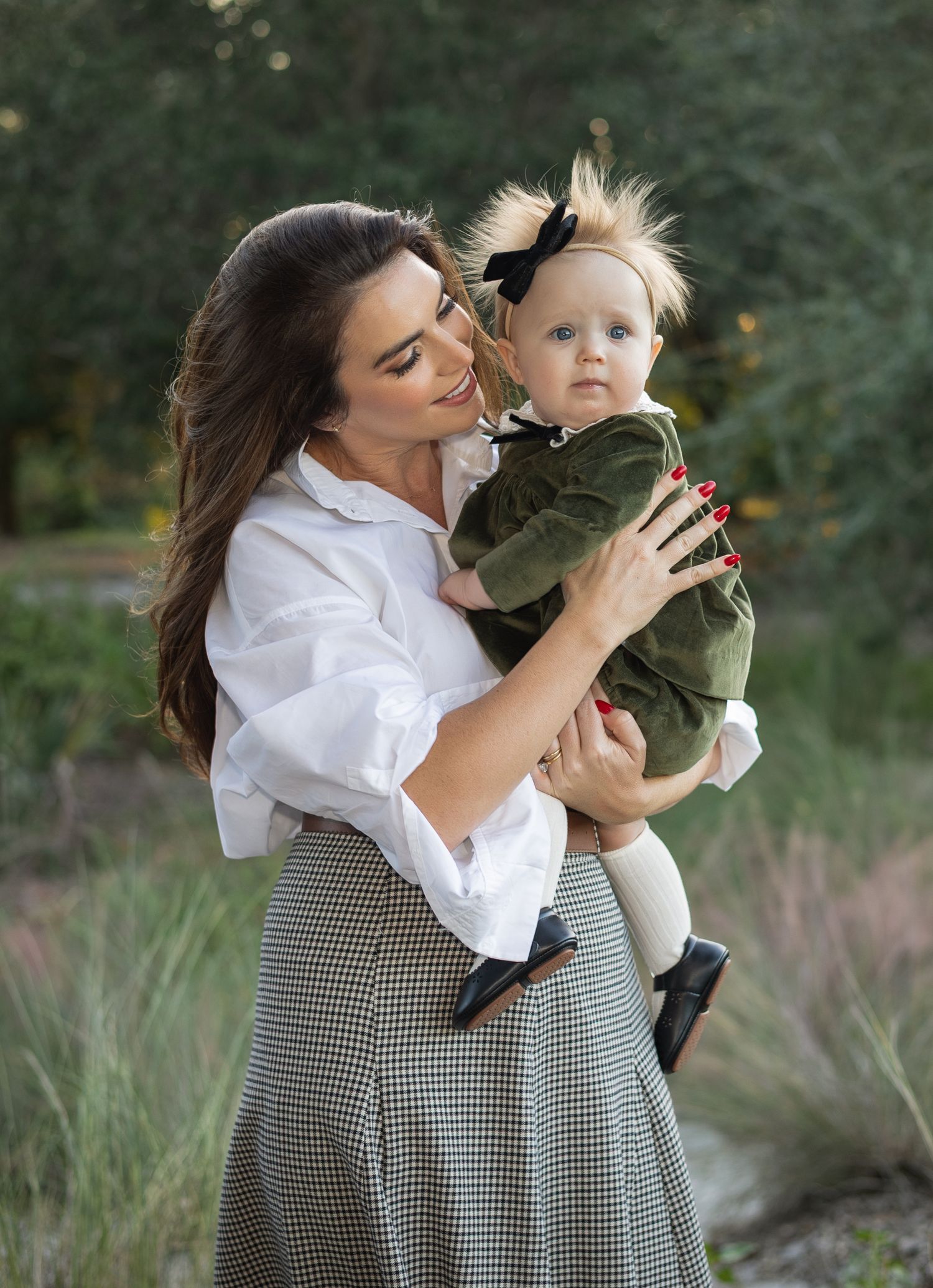 A person in a white blouse and checkered skirt embraces a baby wearing a green outfit in an outdoor setting.