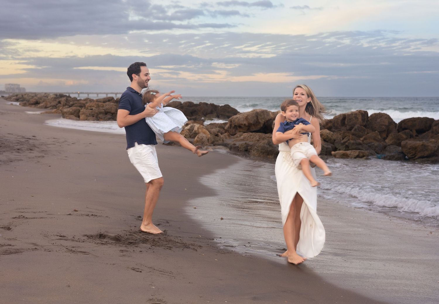 A couple in white and navy outfits dances playfully together on a sandy beach at sunset.