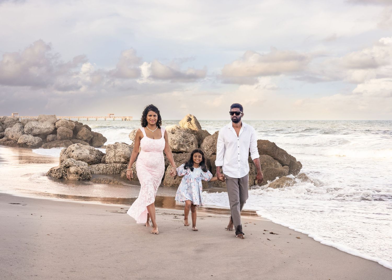 A group in white outfits walks along a scenic beach with crashing waves and rocky outcrops at sunset.