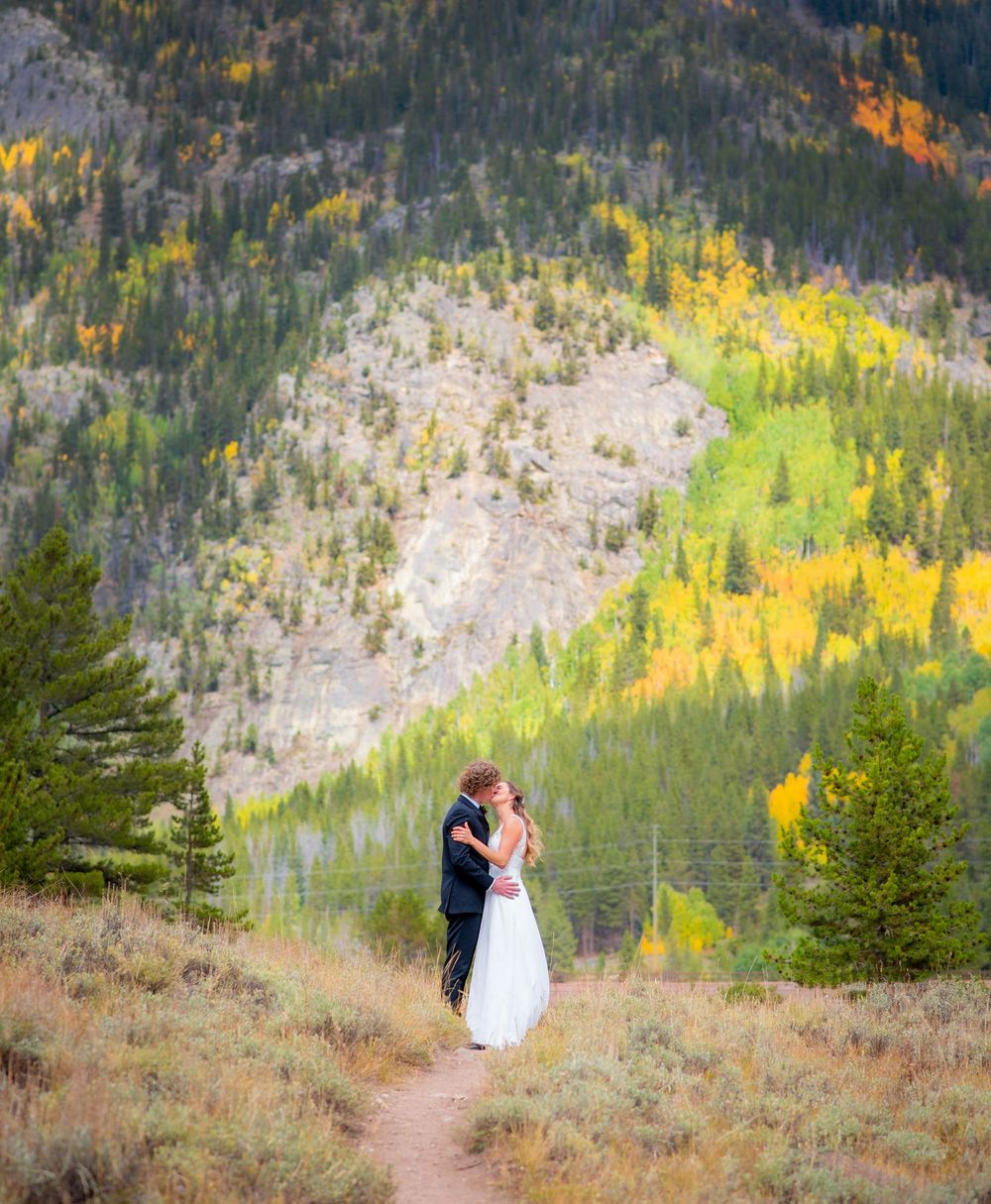 Couple in wedding attire standing on mountain trail with autumn colors in background.