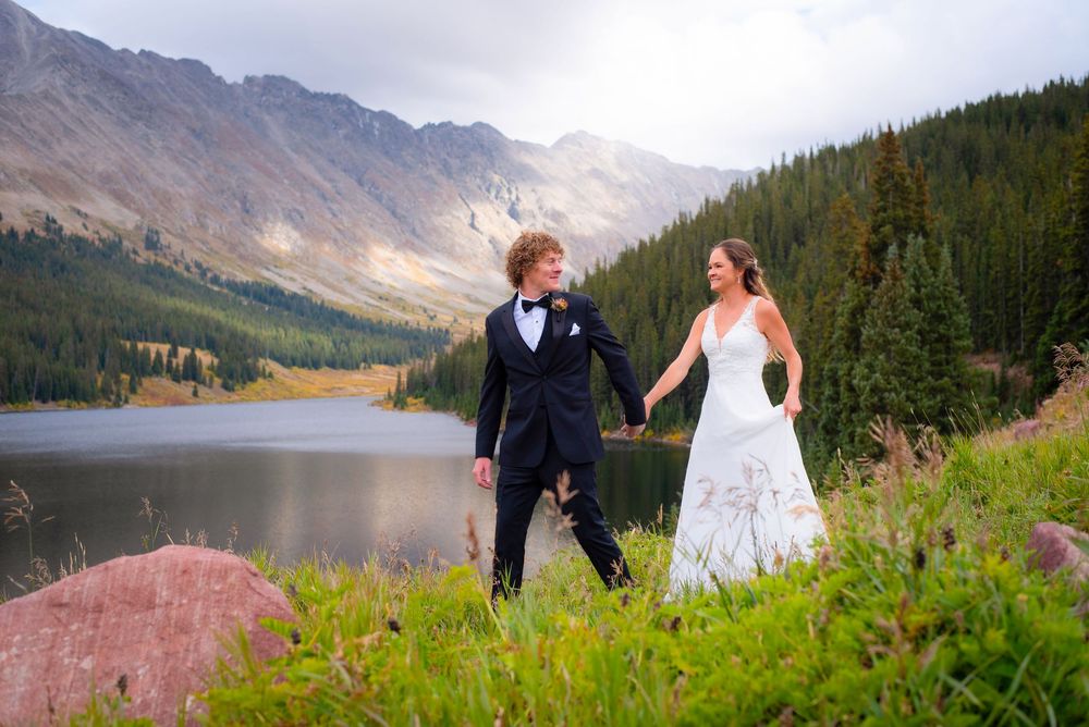 Wedding couple holding hands on hillside with mountain lake and peaks in background.