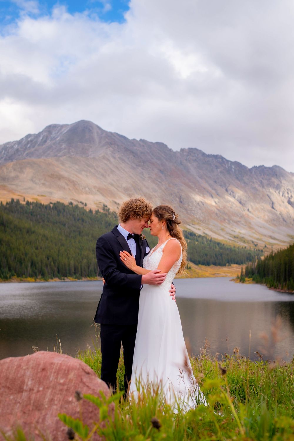 Couple embracing by mountain lake with dramatic peaks and cloudy sky in background.