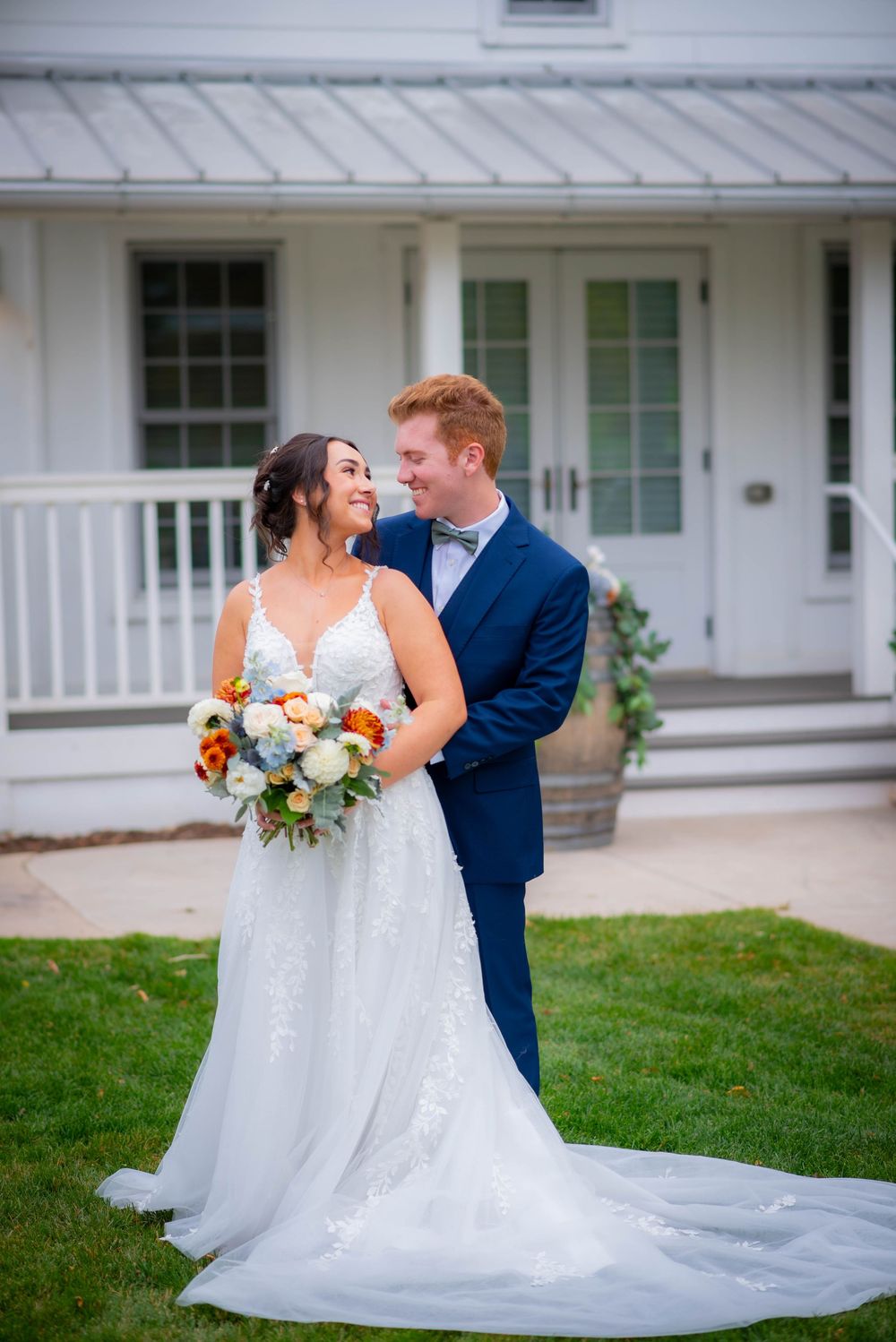Newlyweds pose for portraits outside a white farmhouse with beautiful autumn landscaping.