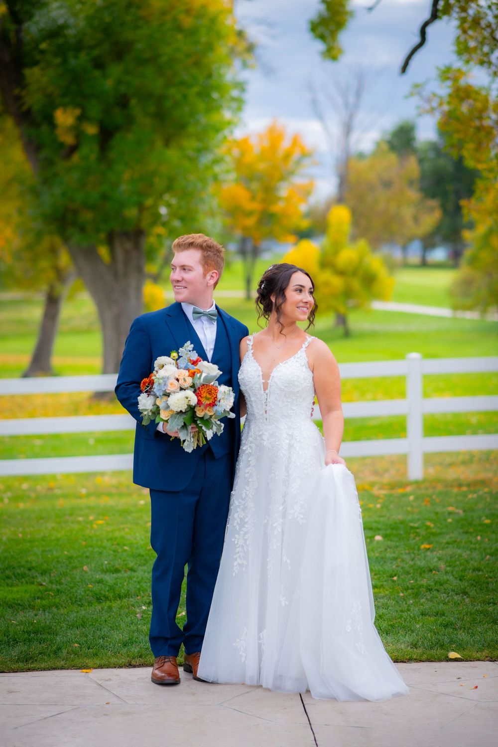 Wedding couple walking together in autumn scenery with white fence and colorful fall foliage.
