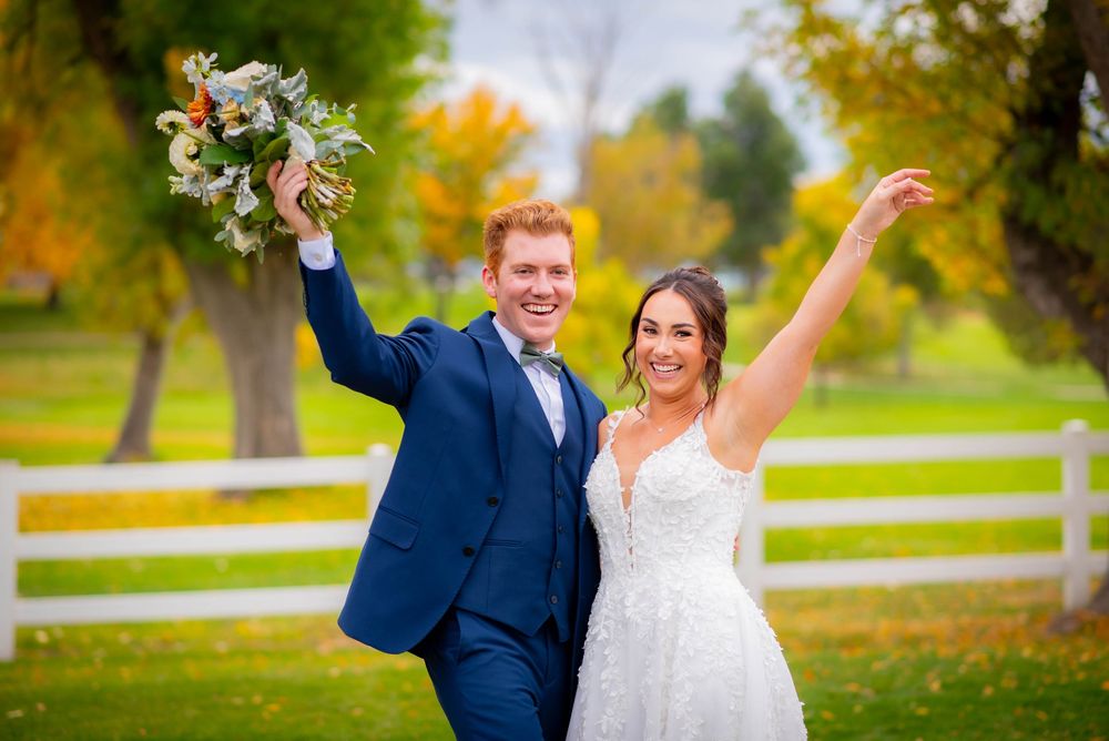 Joyful newlyweds celebrate with raised arms against autumn backdrop with white fence.