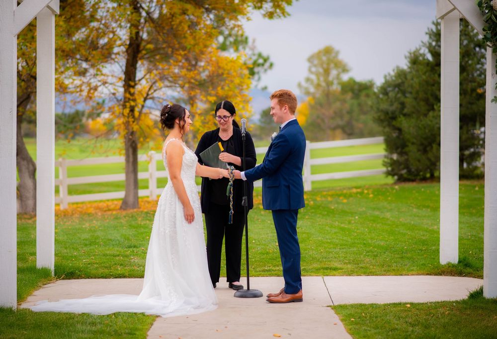 Outdoor wedding ceremony under white pergola with autumn trees and white fence in background.