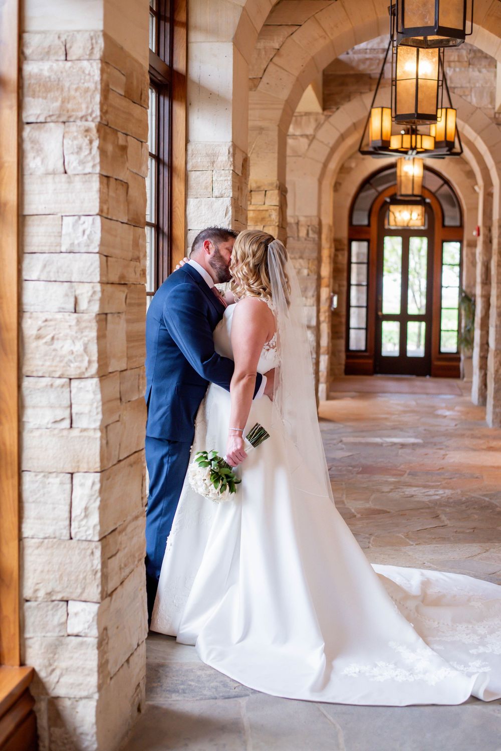 Romantic wedding photo of couple kissing in stone archway with hanging lanterns.