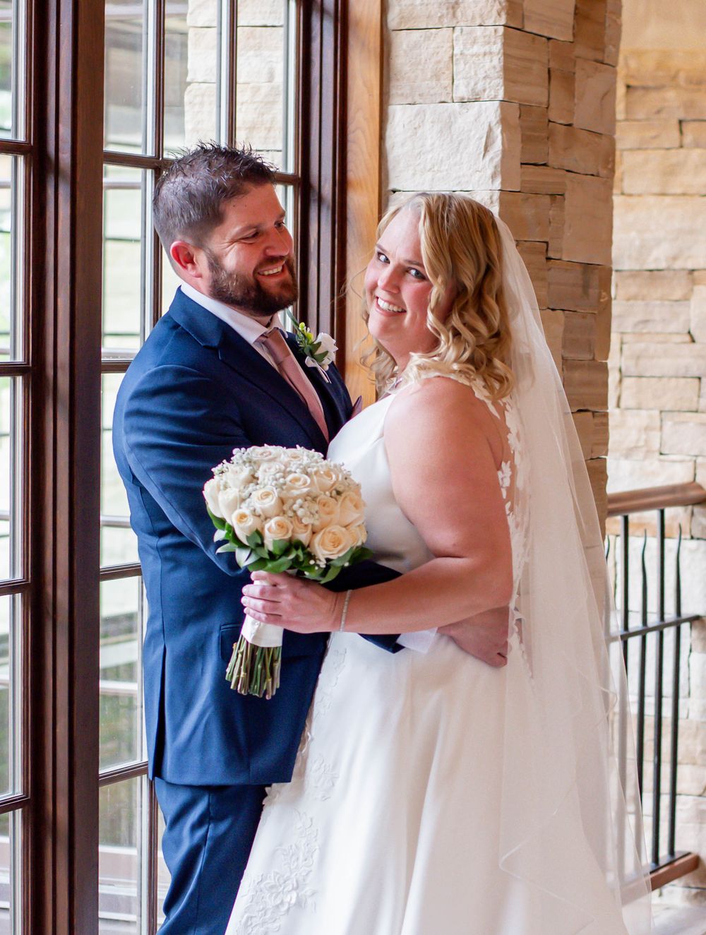 Newlywed couple shares intimate moment while holding white floral bouquet in stone hallway.