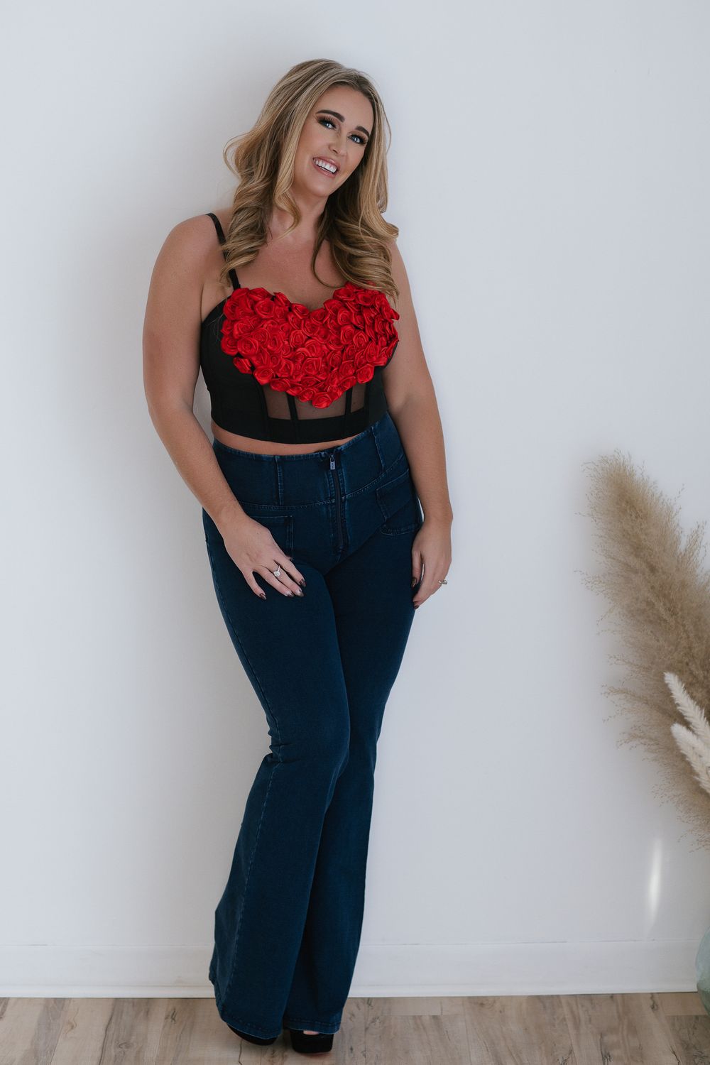 A person wearing a cropped red floral top and high-waisted dark blue jeans poses against a white wall with pampas grass decor.