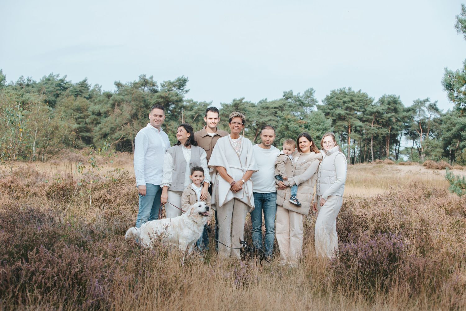 A group wearing neutral toned clothing poses together with a dog in a heathland landscape with pine trees in the background.