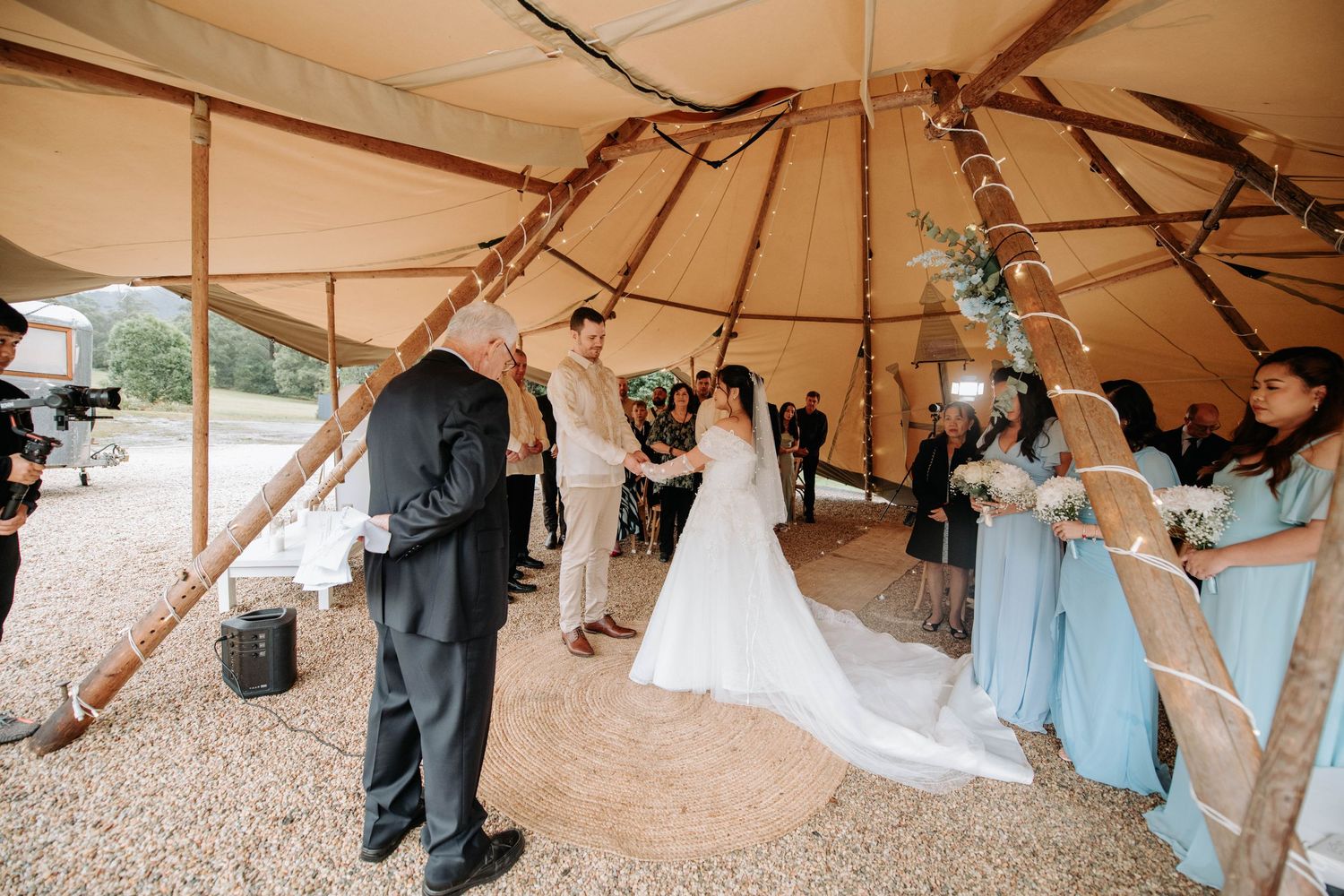 Wedding ceremony taking place inside a rustic teepee tent with wooden poles.