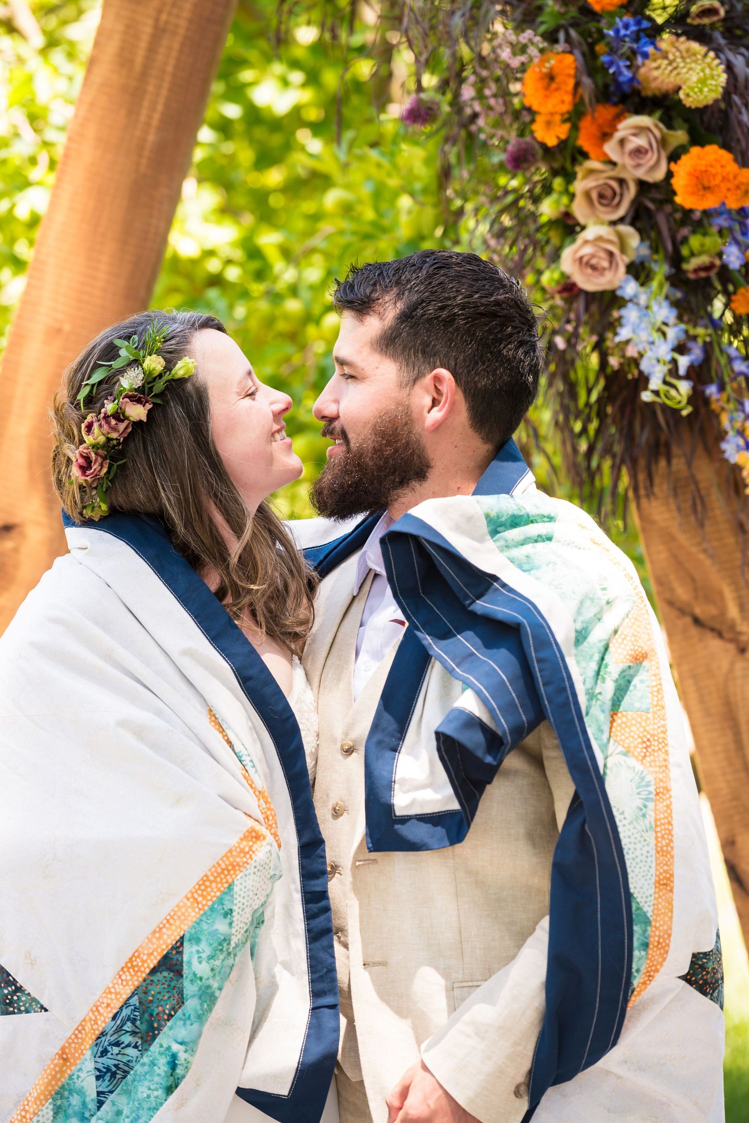 A couple shares an intimate moment under floral decorations wearing unique patterned shawls.