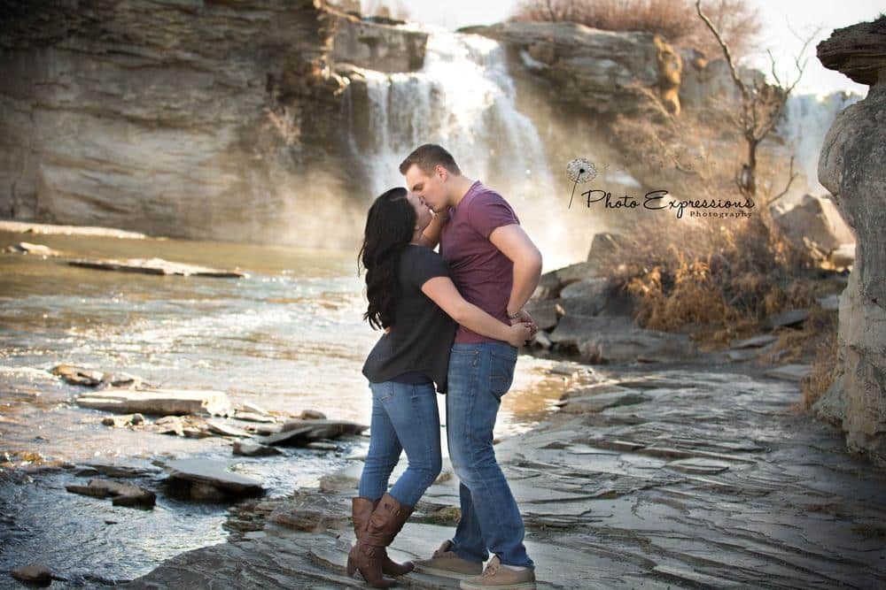 Couple embracing near rocky waterfall during outdoor photo session.