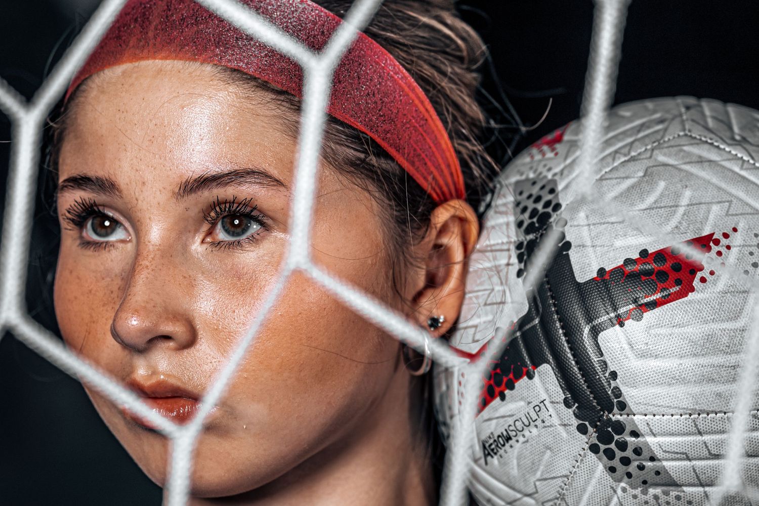A female soccer player is photographed through the net of a goal and has a soccer ball near to her head.