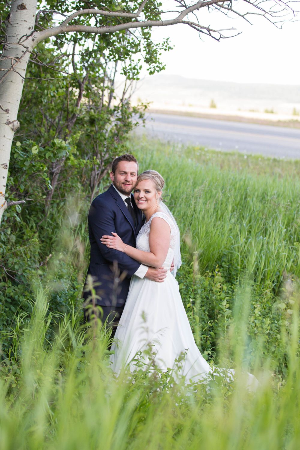 Newlyweds embracing in a field of tall grass with white birch trees in the background.