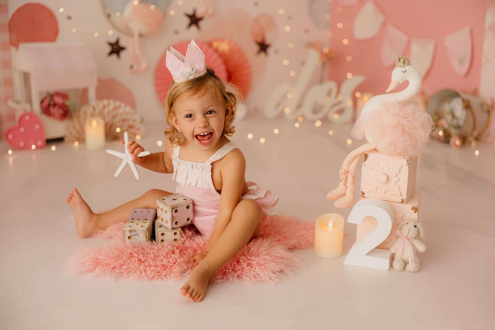 Fotografía del segundo cumpleaños de una niña sonriendo con una corona y peto vichy rosa con decoración en tonos rosas.