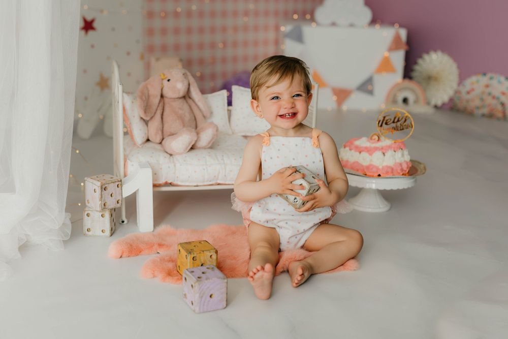 Fotografía del primer cumpleaños de una niña con tarta y peto en tonos pastel con decoración en rosa y lila