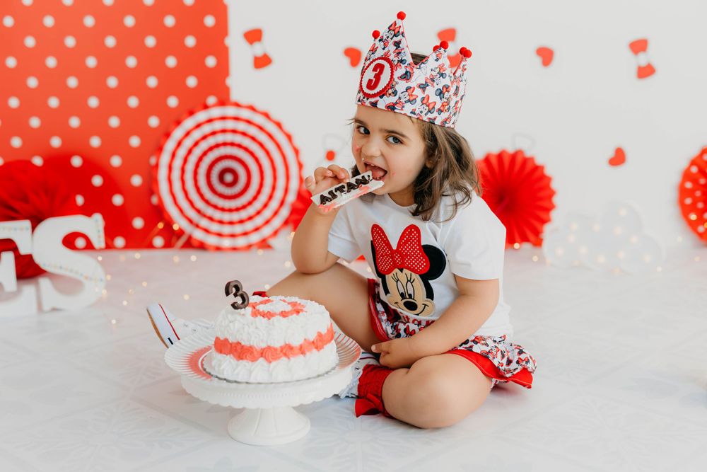 Fotografía del tercer año de una niña comiendo tarta con decoración en tonos rojos en su sesión en estudio.
