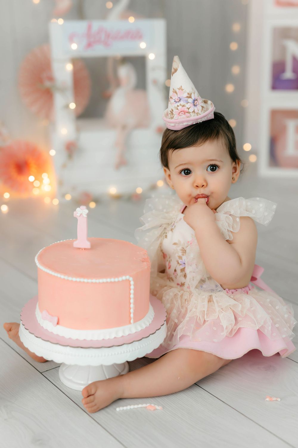Fotografía del primer cumpleaños de una niña comiendo tarta con un cono y traje floral, con decoración en tonos rosas