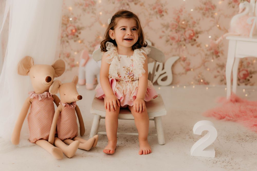 Fotografía del segundo cumpleaños de una niña sonriendo con un peto floral  y decoración en tonos rosas